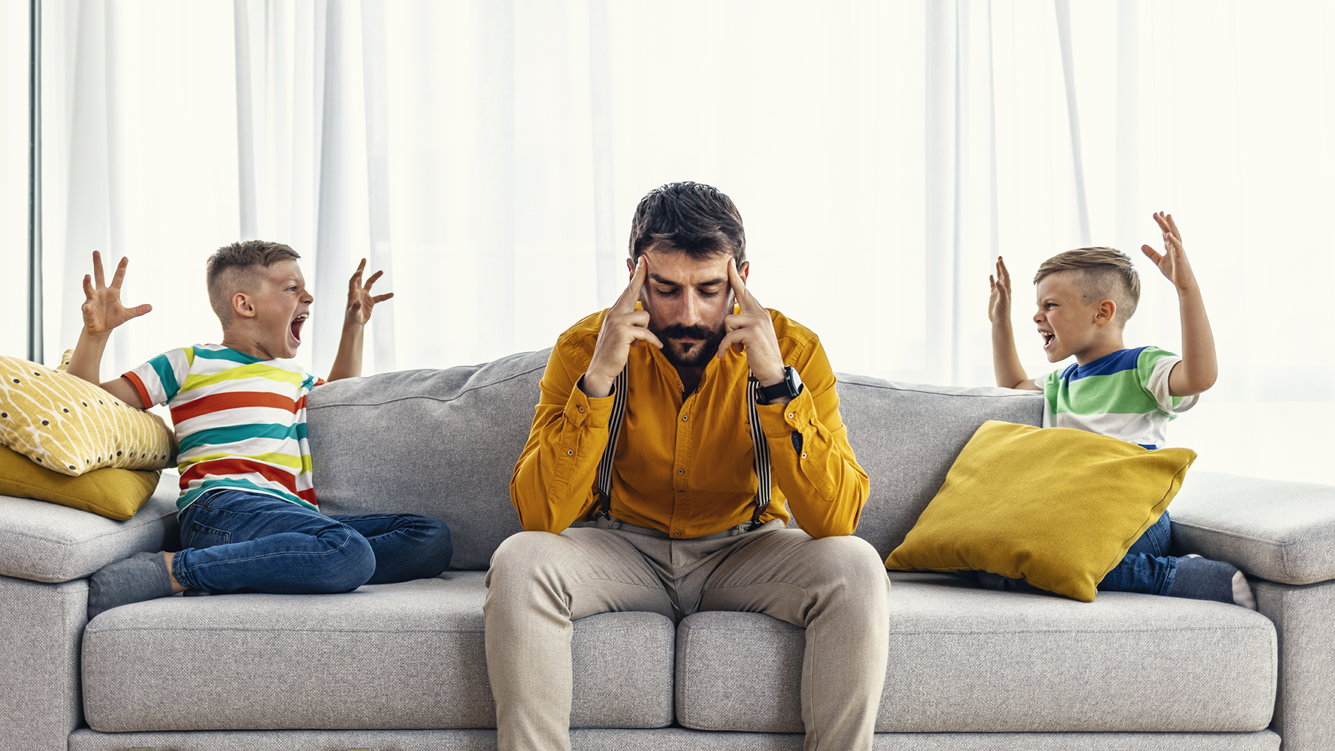 Isä ja kaksi lasta sohvalla. Lapset näyttävät riitelevän ja isä sulkee korviansa käsillään.