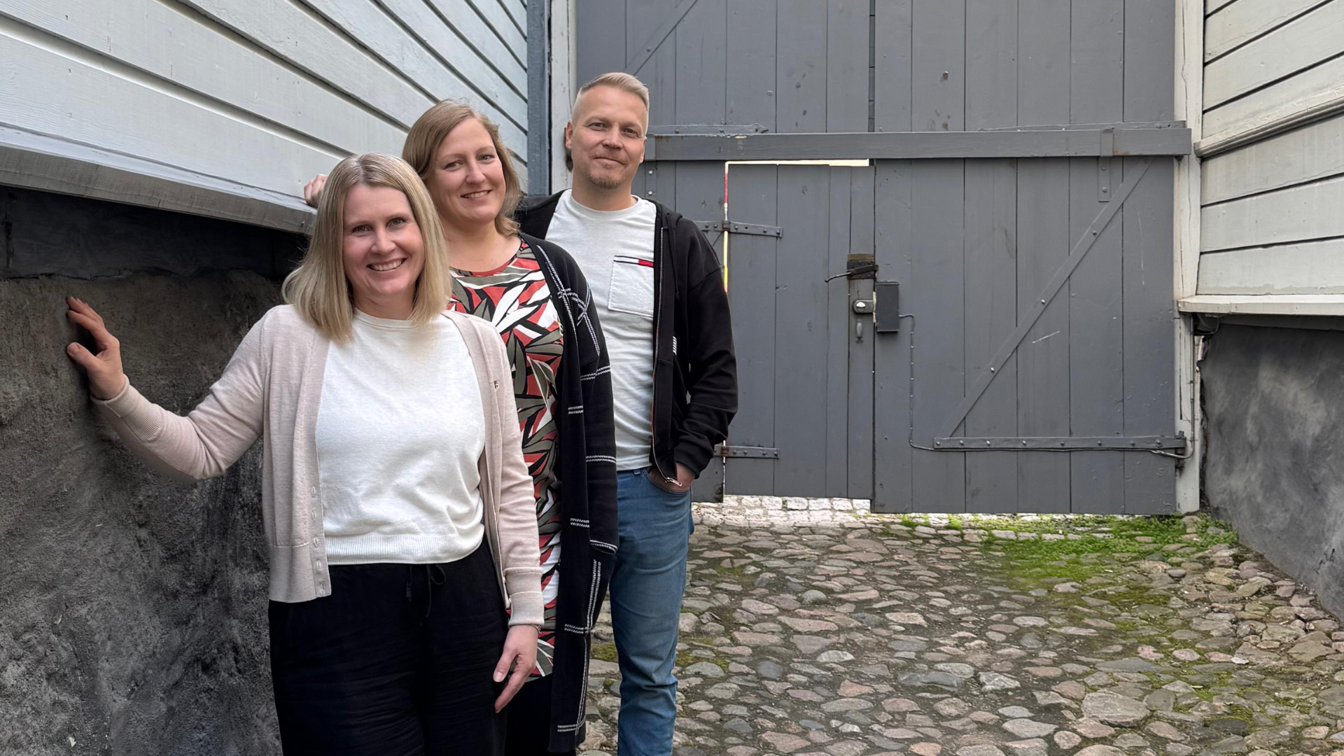 Kolme henkilöä seisoo ja hymyilee valoisassa porttikongissa. Takana iso puuportti ja reunoilla korkeat kivijalat, josta kohoavat vanhat puutalot. Mukulakivimaa.