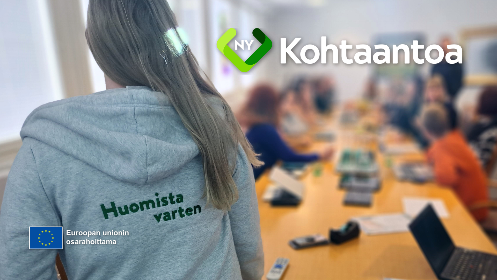 Etualalla henkilö harmaassa hupparissa, jossa lukee 'Huomista varten'. Taustalla pitkä pöytä, jonka ympärillä istuu ryhmä ihmisiä kokouksessa. Kuvan yläosassa näkyy NY Kohtaantoa -logo sekä Euroopan unionin osarahoittama -merkintä.
