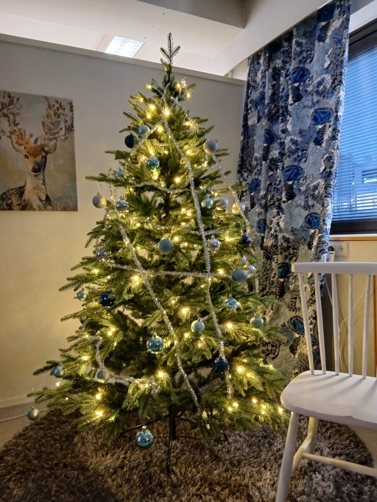 Kuva koristellusta joulukuusesta hämyisessä tunnelmallisessa valaistuksessa. Kuusessa lämpimän valkoiset valot, kylmän sinisen sävyisiä kuusen palloja ja hopeisia ohuita nauhoja.