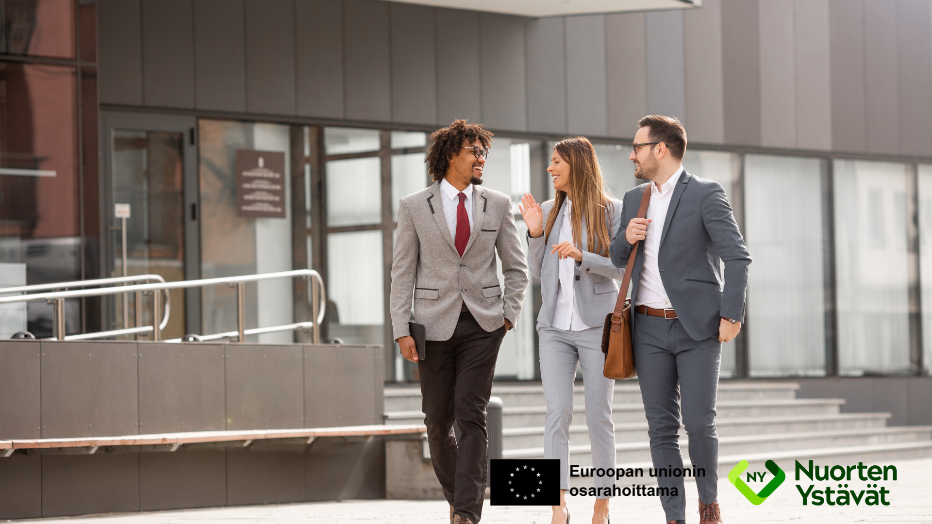 Kolme pukuihin pukeutunutta henkilöä kävelee keskustellen modernin rakennuksen edessä.